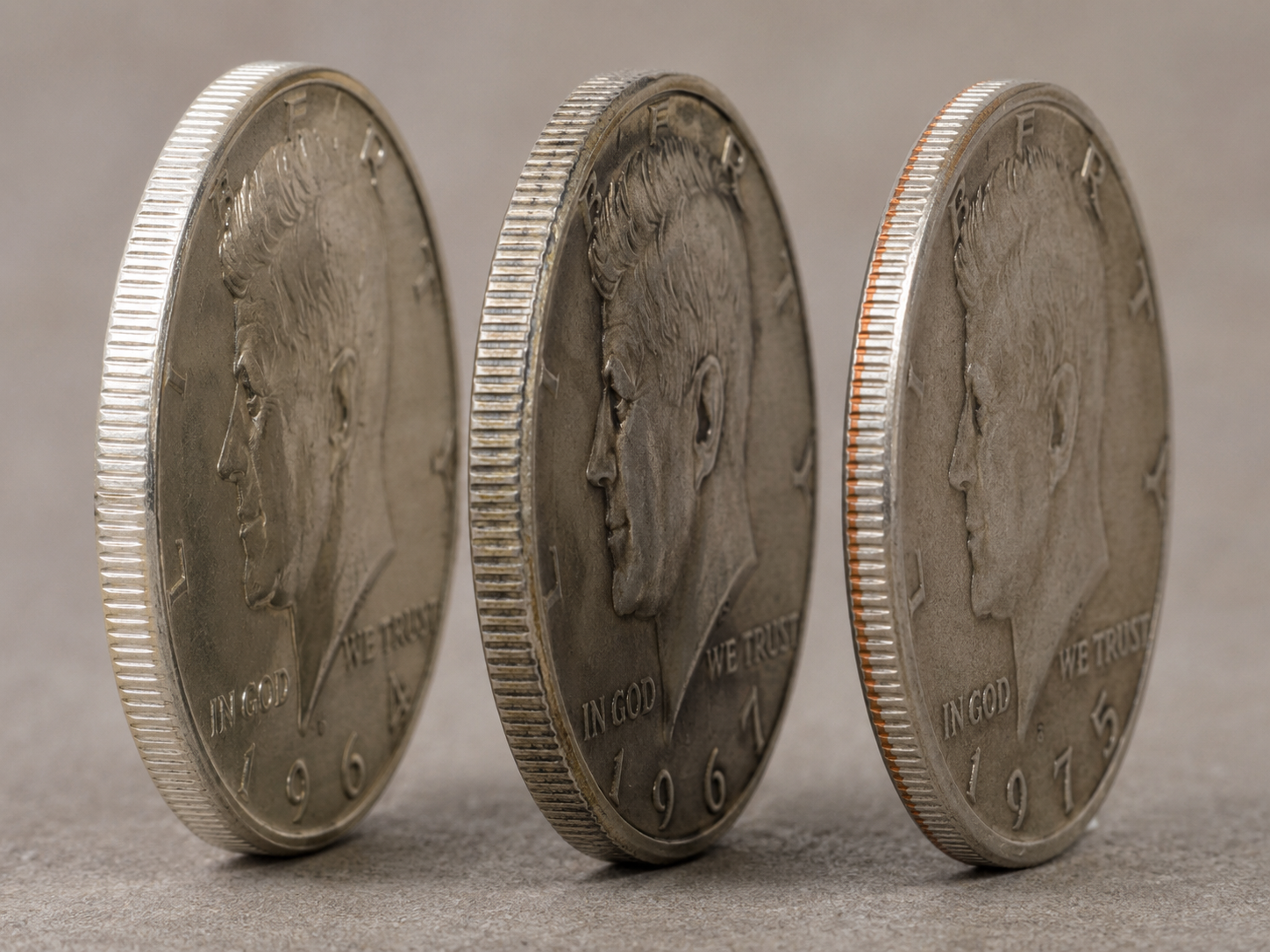 Three Kennedy half dollars on edge — 1964 90% silver, 1967 40% silver, and 1975 clad — showing the progressive copper tinge across compositions.