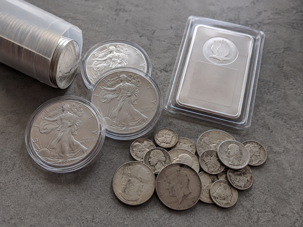A small silver stack: 1 oz American Silver Eagles in capsules, a 10 oz silver bar, and a pile of pre-1965 90% silver U.S. coins.